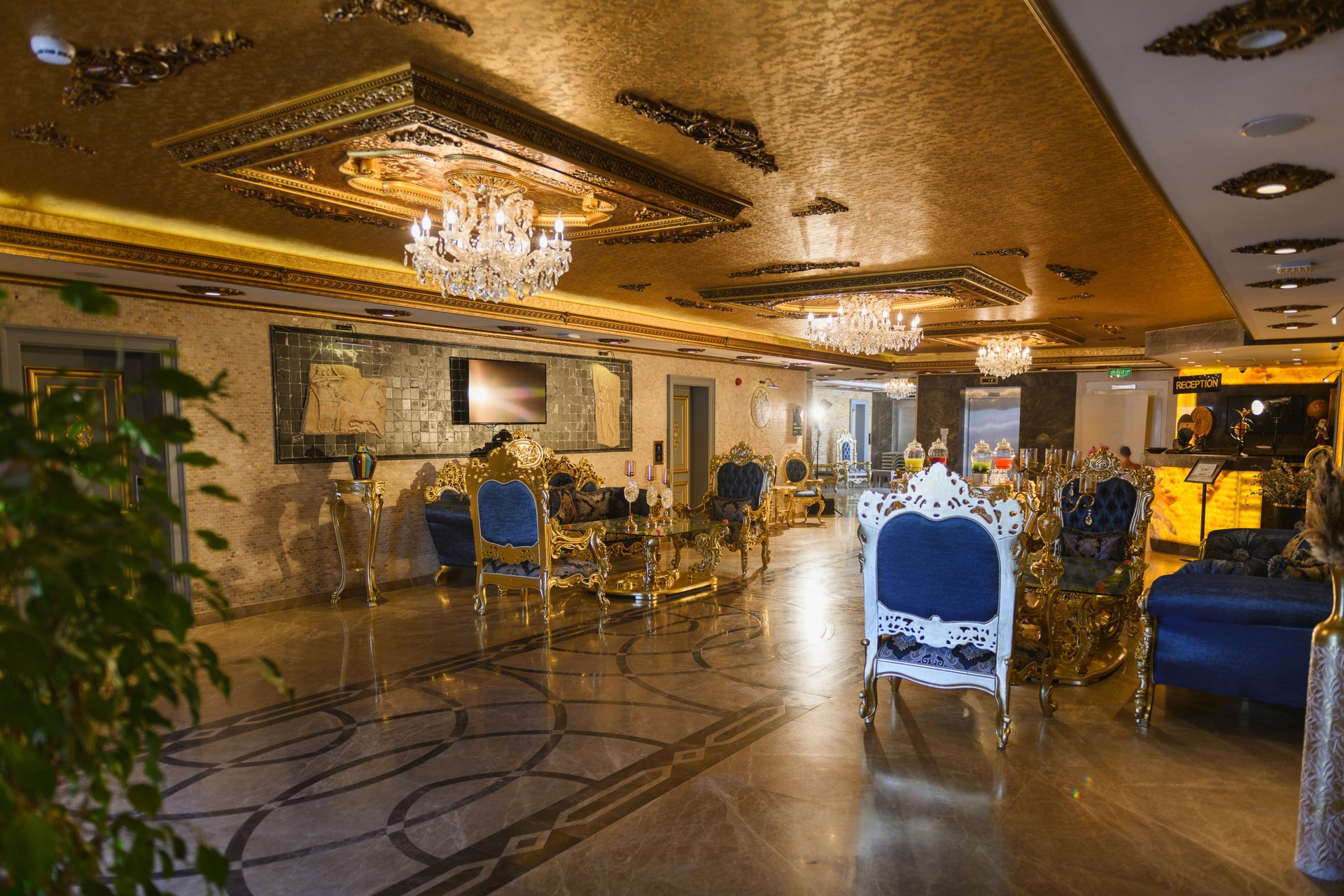 Opulent hotel lobby in baroque style with gold accents, blue velvet armchairs and sofas, marble floors, grand chandeliers, and wall-mounted televisions